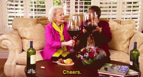 Two people sit on a couch clinking oversized wine glasses together, with bottles of wine and food on the table in front of them. The word Cheers. appears at the bottom of the image.